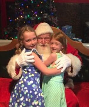 Two girls happily hugging Santa Claus indoors.